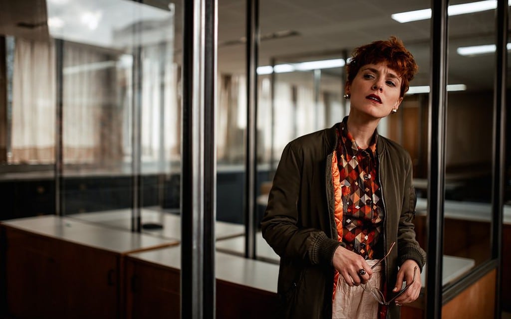 Una mujer elegante con cabello corto rojo piensa en un espacio de oficina, apoyándose ligeramente contra una partición de vidrio.